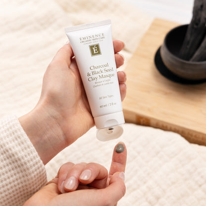 Hand holding a tube of Eminence Charcoal & Black Seed Clay Masque on a neutral background